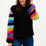 A woman wearing a black sweater with ribbed cuffs and vibrant rainbow-colored sleeves, paired with blue jeans.
