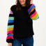 A woman wearing a black sweater with ribbed cuffs and vibrant rainbow-colored sleeves, paired with blue jeans.