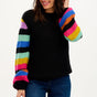 A woman wearing a black sweater with ribbed cuffs and vibrant rainbow-colored sleeves, paired with blue jeans.
