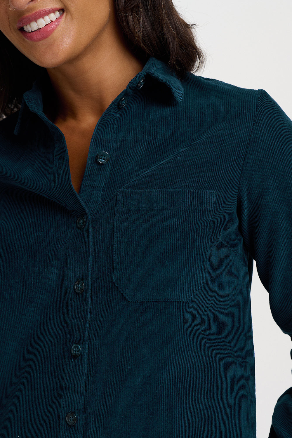 Close-up of a woman wearing a dark teal corduroy button-up shirt with a chest pocket, against a light gray background.