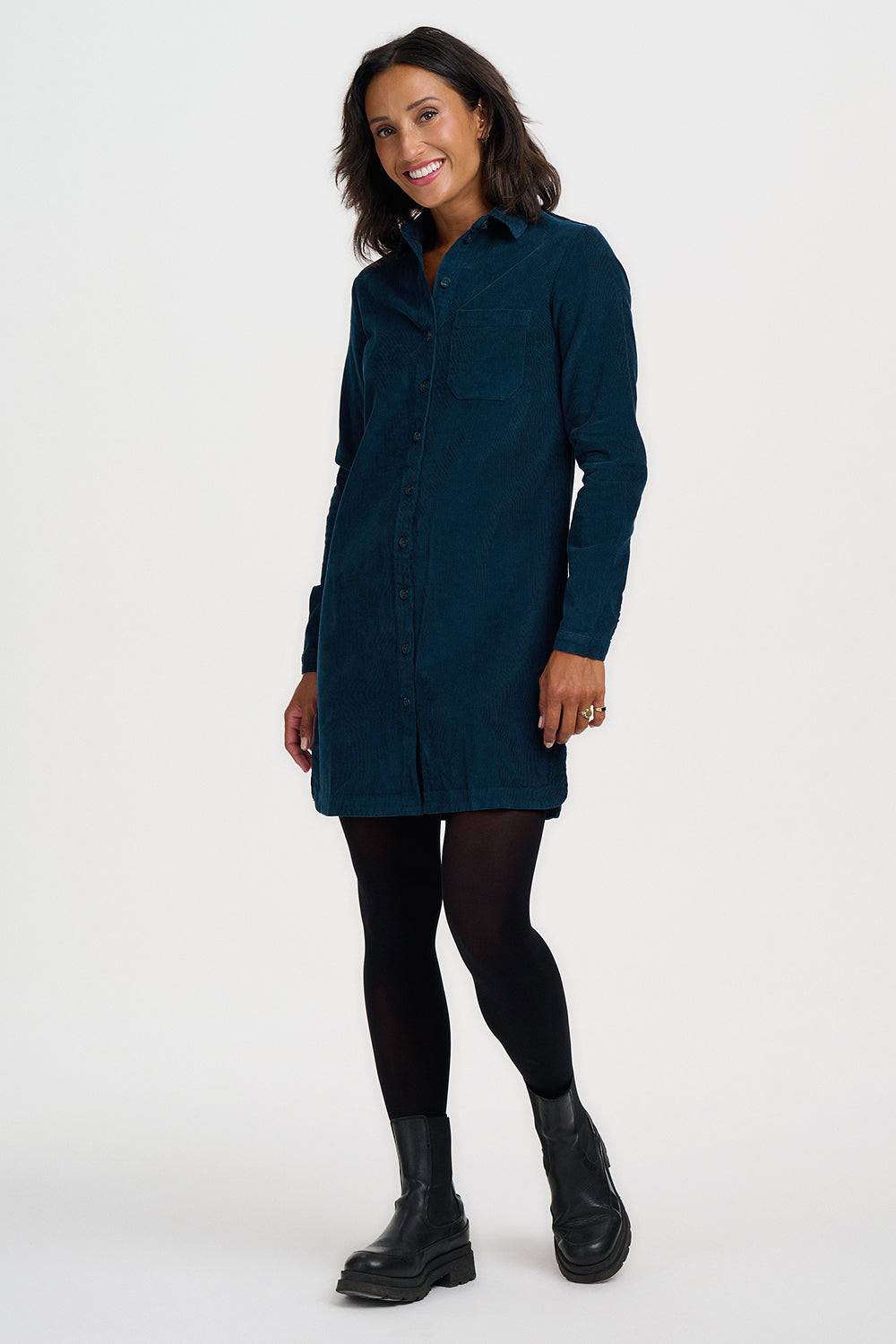 A woman modeling a dark teal corduroy shirt dress with black tights and black leather ankle boots, standing against a plain white studio background.