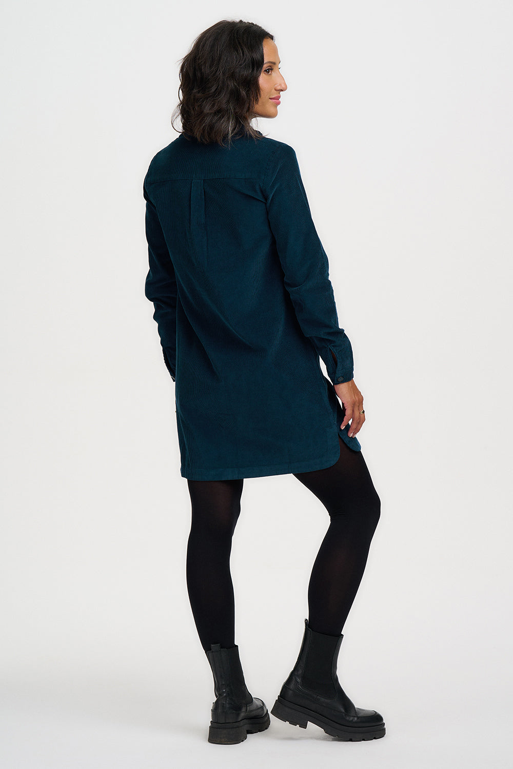 A woman wearing a dark teal corduroy shirt dress, styled with black tights and black platform boots, shown from a three-quarters back view against a plain white studio background.