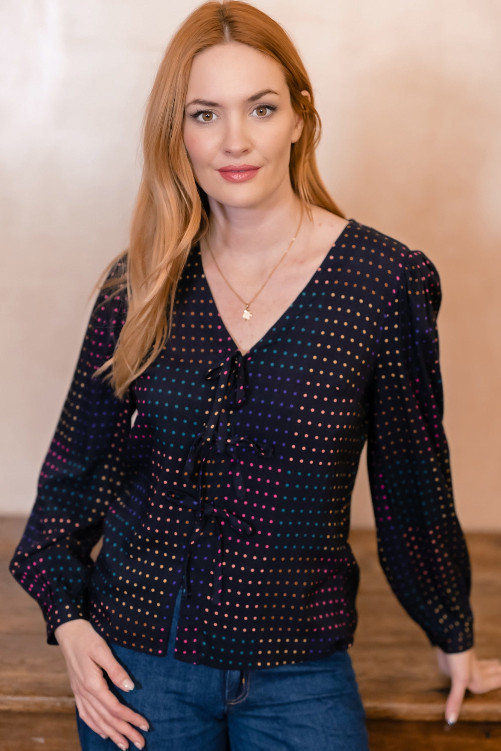 A woman with long red hair wearing a dark navy long-sleeve blouse with a colorful polka dot pattern and blue jeans, standing against a warm beige background.