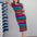 Woman wearing a colorful striped dress standing next to a rack of shoes against a plain wall.