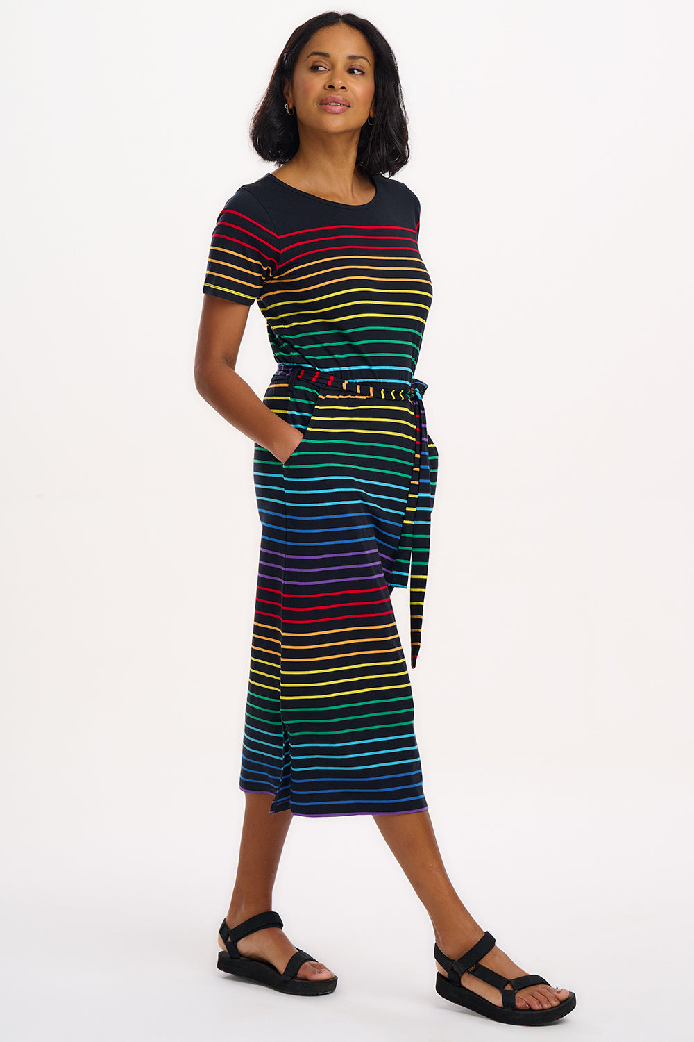 A model wearing a midi-length jersey dress with a black background featuring a vibrant multicolour striped pattern. The dress has a t-shirt fit with a matching tie belt and short sleeves.