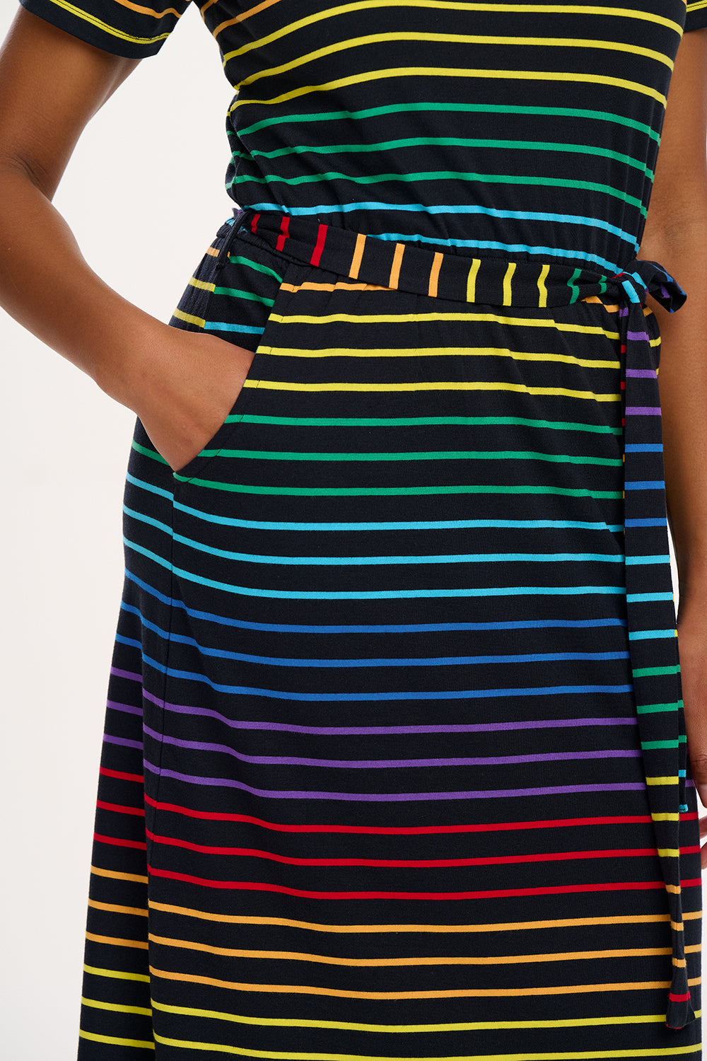 A model wearing a midi-length jersey dress with a black background featuring a vibrant multicolour striped pattern. The dress has a t-shirt fit with a matching tie belt and short sleeves.