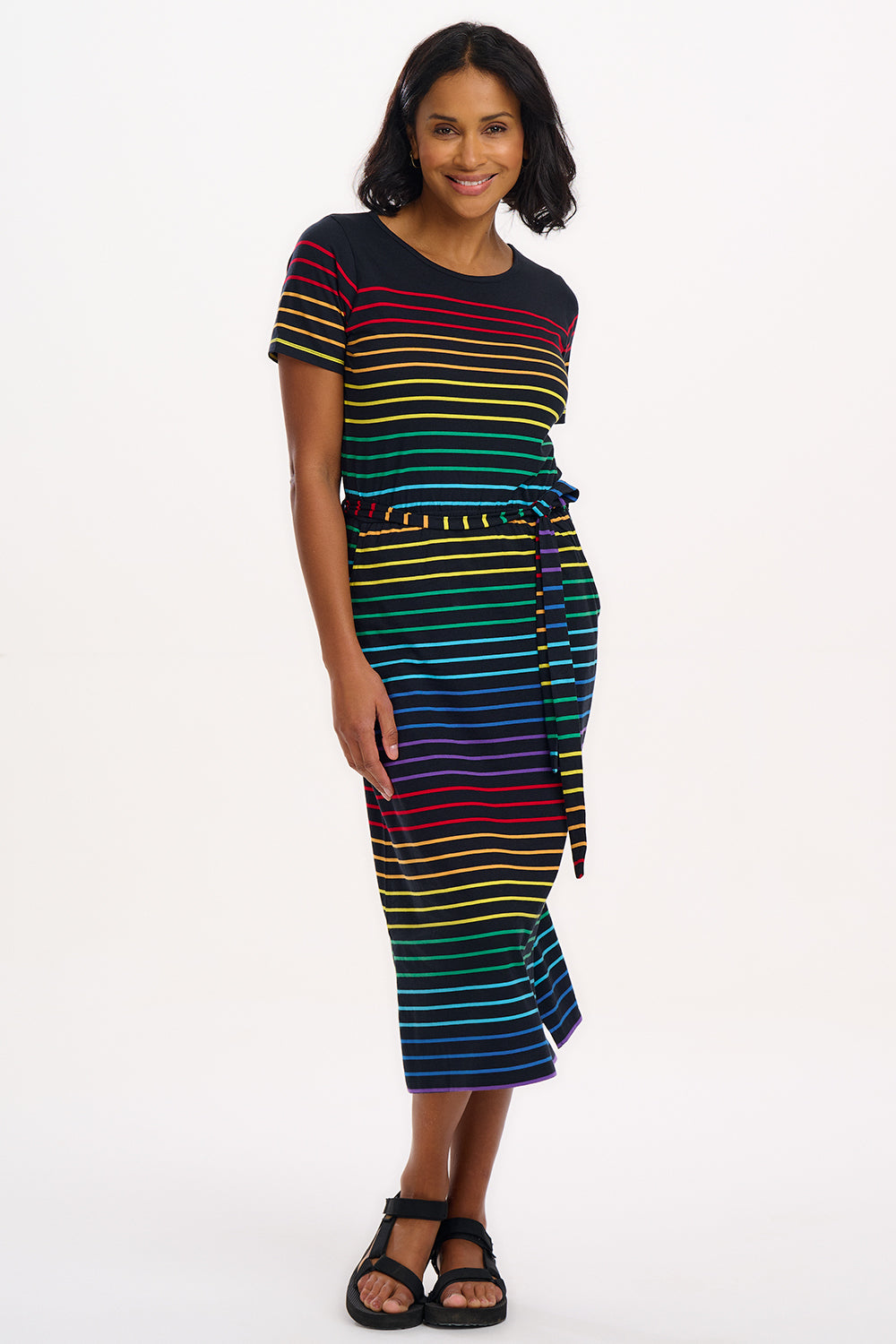 A model wearing a midi-length jersey dress with a black background featuring a vibrant multicolour striped pattern. The dress has a t-shirt fit with a matching tie belt and short sleeves.