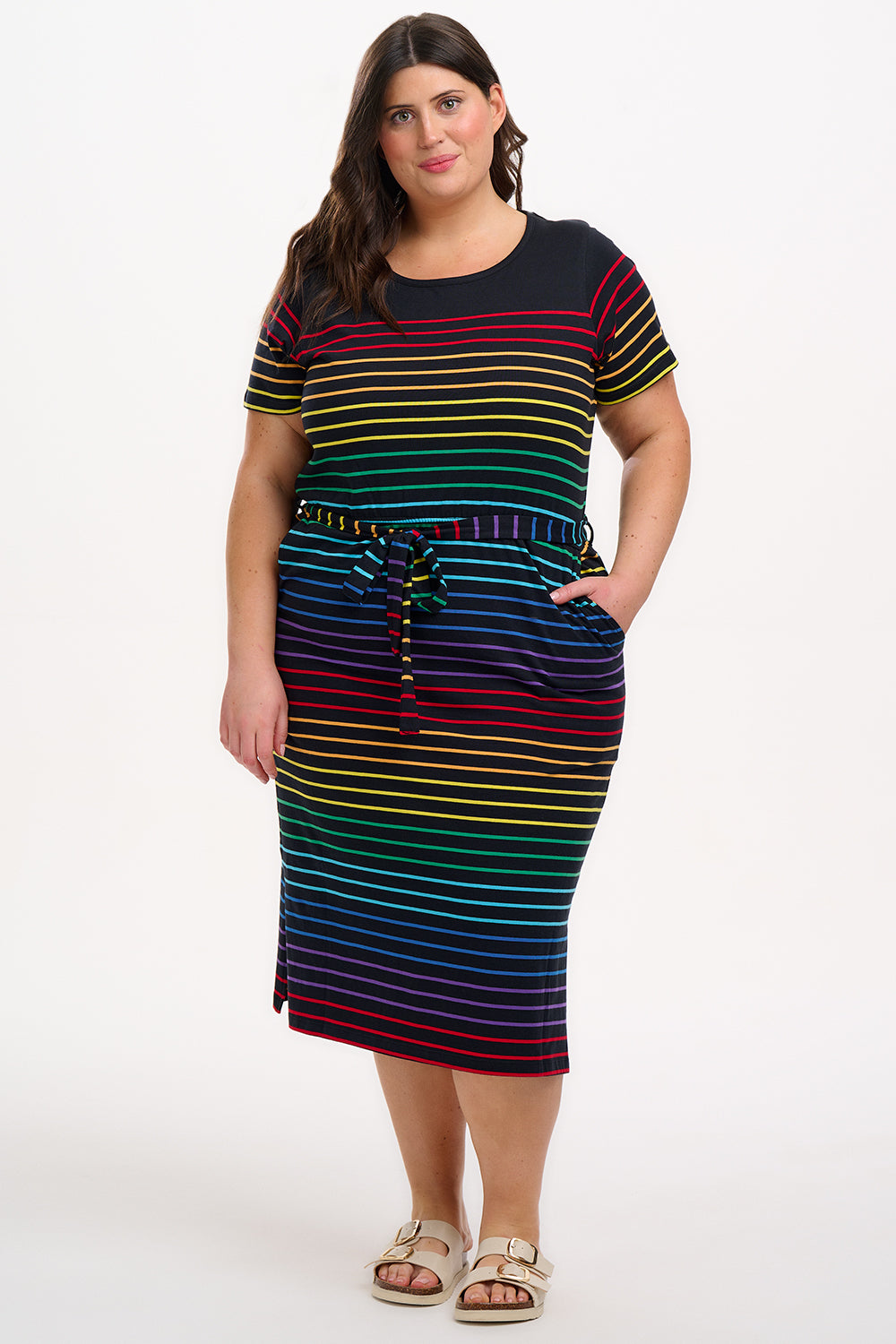 A model wearing a midi-length jersey dress with a black background featuring a vibrant multicolour striped pattern. The dress has a t-shirt fit with a matching tie belt and short sleeves.