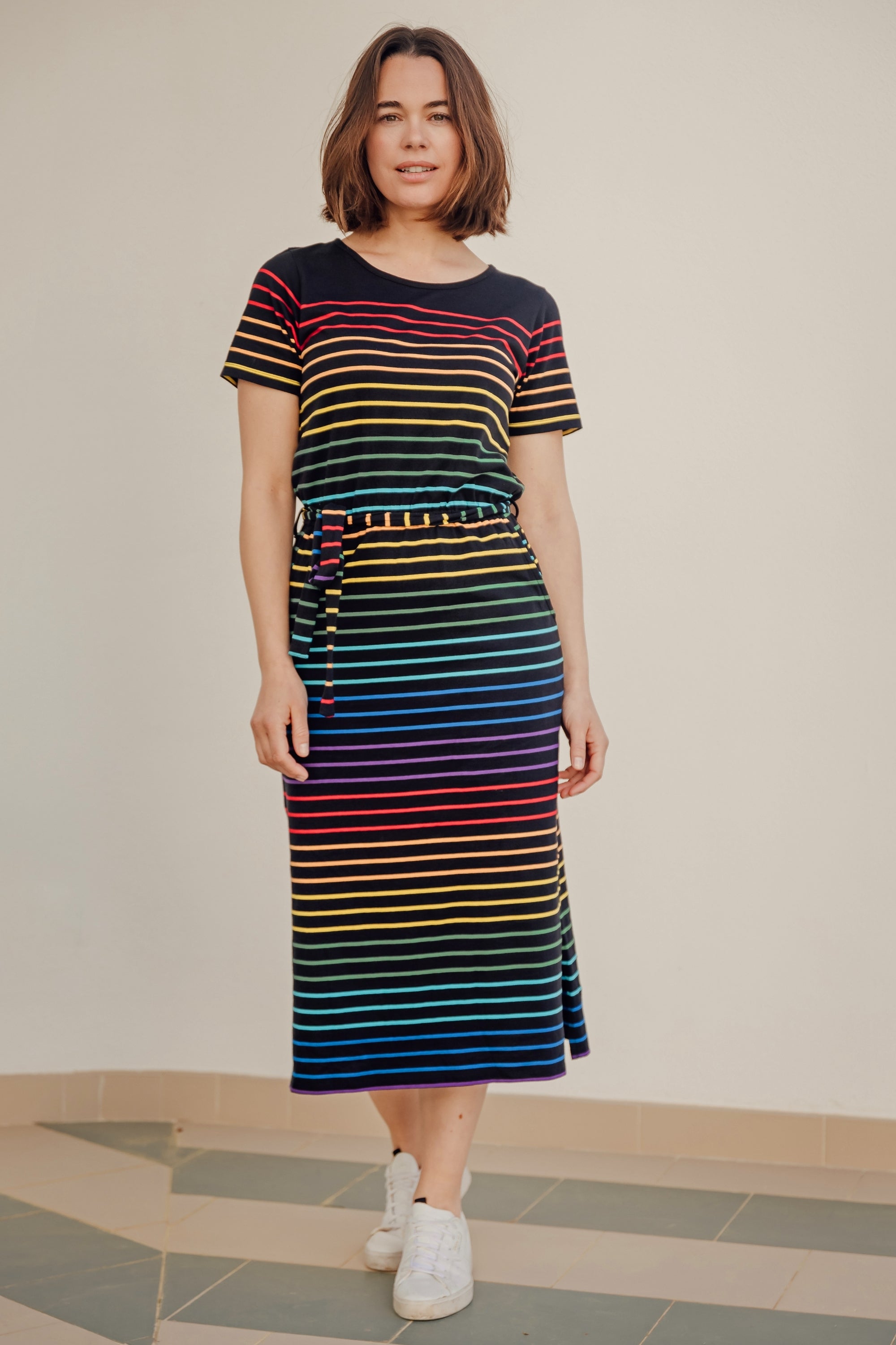 A model wearing a midi-length jersey dress with a black background featuring a vibrant multicolour striped pattern. The dress has a t-shirt fit with a matching tie belt and short sleeves.