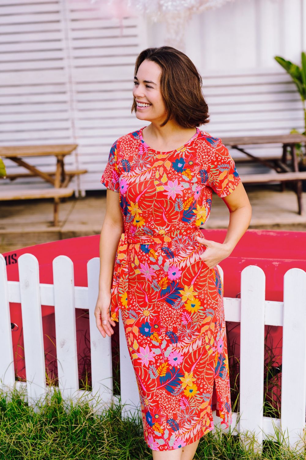 Woman wearing a colorful floral dress standing in front of a white picket fence.