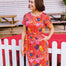 Woman wearing a colorful floral dress standing in front of a white picket fence.