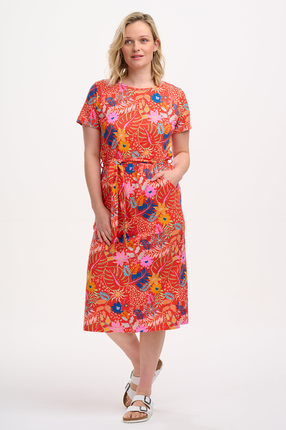 A model wearing a red jersey midi dress with a colorful tropical leaf and flower print, standing against a plain background.