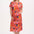 A model wearing a red jersey midi dress with a colorful tropical leaf and flower print, standing against a plain background.