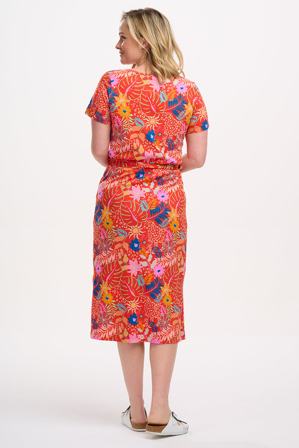 A model wearing a red jersey midi dress with a colorful tropical leaf and flower print, standing against a plain background.