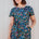 A person wearing a teal and multicolored jersey midi dress with a floral and dinosaur pattern, featuring a t-shirt fit upper, matching tie belt, and pockets on a midi length skirt.