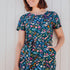 A person wearing a teal and multicolored jersey midi dress with a floral and dinosaur pattern, featuring a t-shirt fit upper, matching tie belt, and pockets on a midi length skirt.