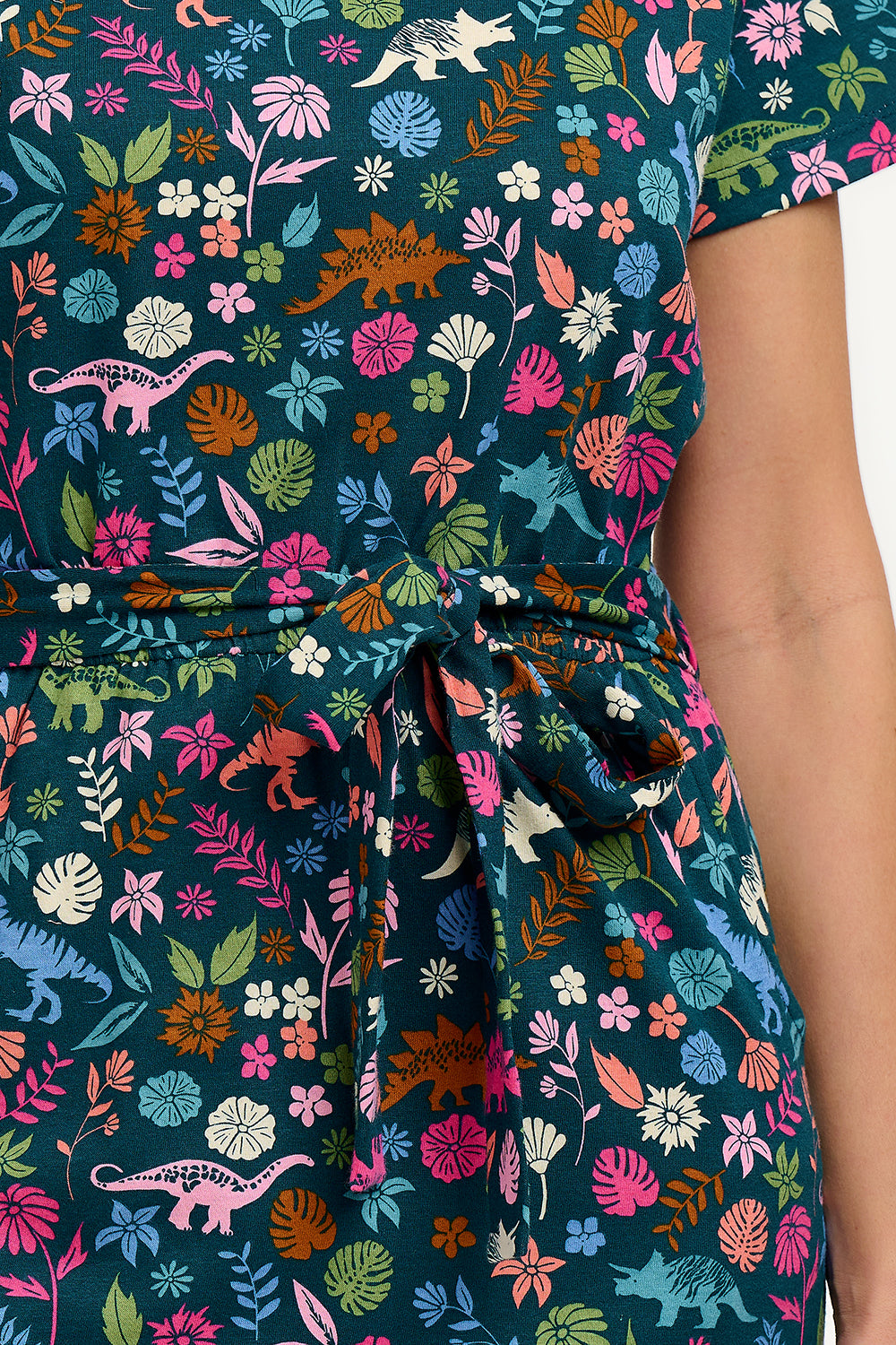 A model wearing a mid-length jersey dress with a colorful dinosaur and flower print, in teal and multicolor, with a matching tie belt.