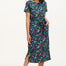 A model wearing a mid-length jersey dress with a colorful dinosaur and flower print, in teal and multicolor, with a matching tie belt.