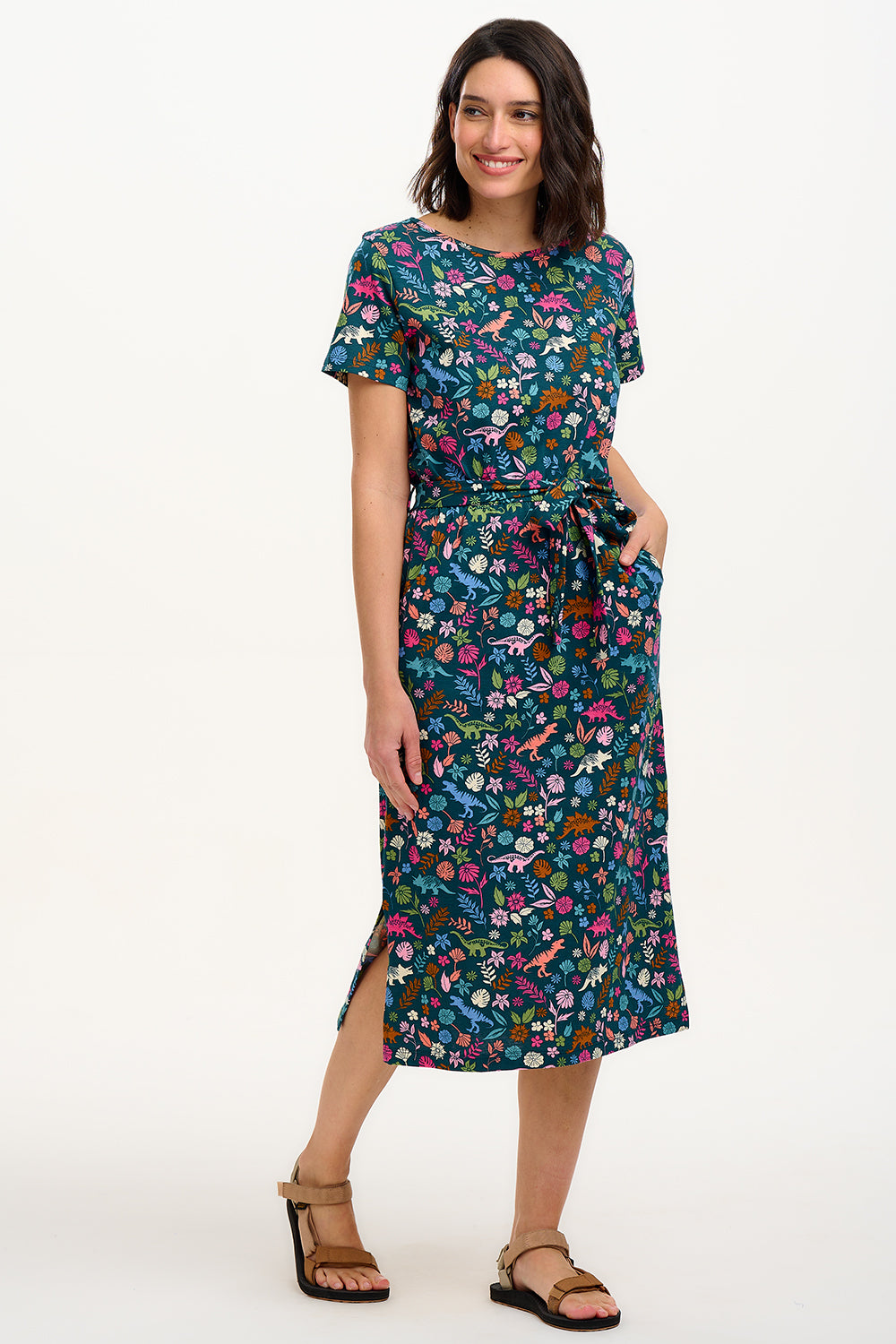 A model wearing a mid-length jersey dress with a colorful dinosaur and flower print, in teal and multicolor, with a matching tie belt.