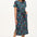 A model wearing a mid-length jersey dress with a colorful dinosaur and flower print, in teal and multicolor, with a matching tie belt.