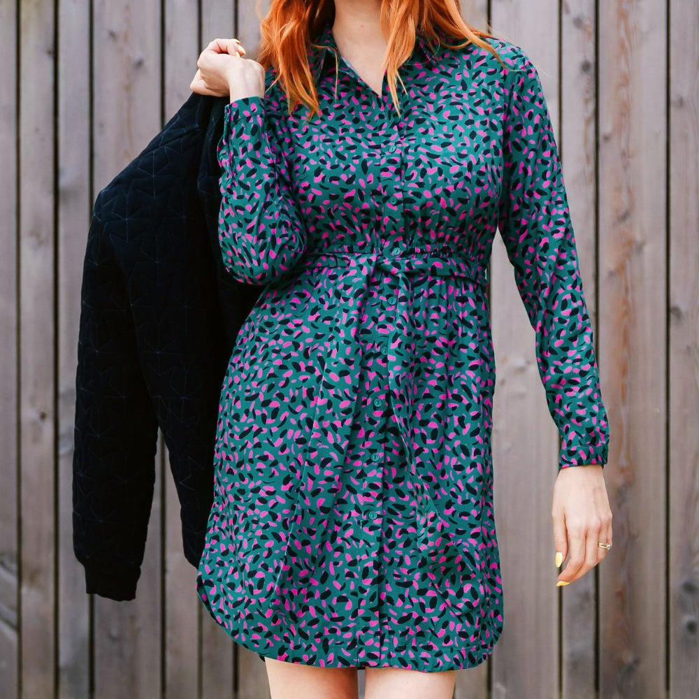 Woman wearing a patterned dress holding a black jacket against a wooden fence background