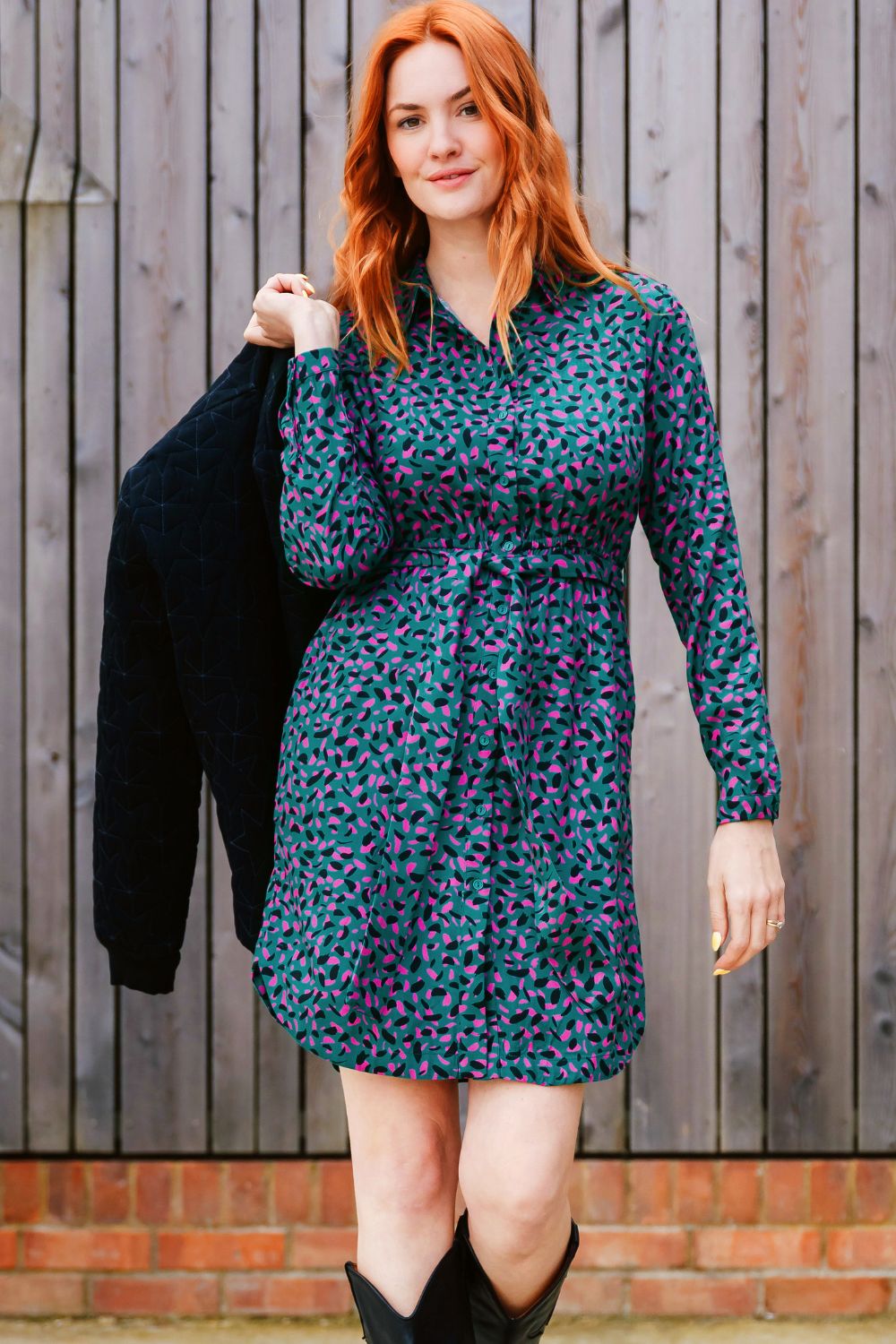 Woman wearing a patterned dress holding a black jacket against a wooden fence background