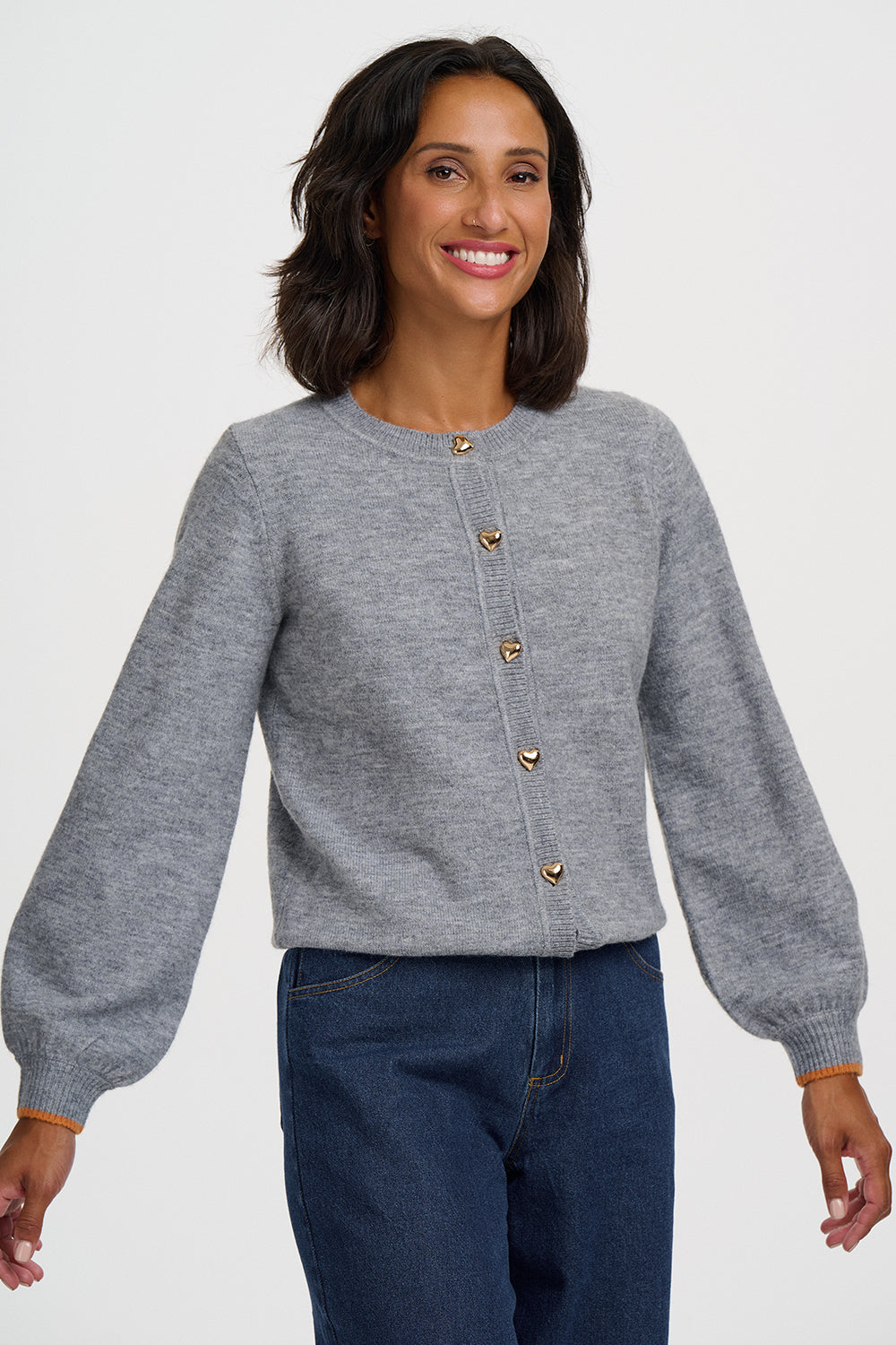 A woman wearing a light gray cropped cardigan with gold buttons and dark blue jeans, photographed against a plain light gray studio background.