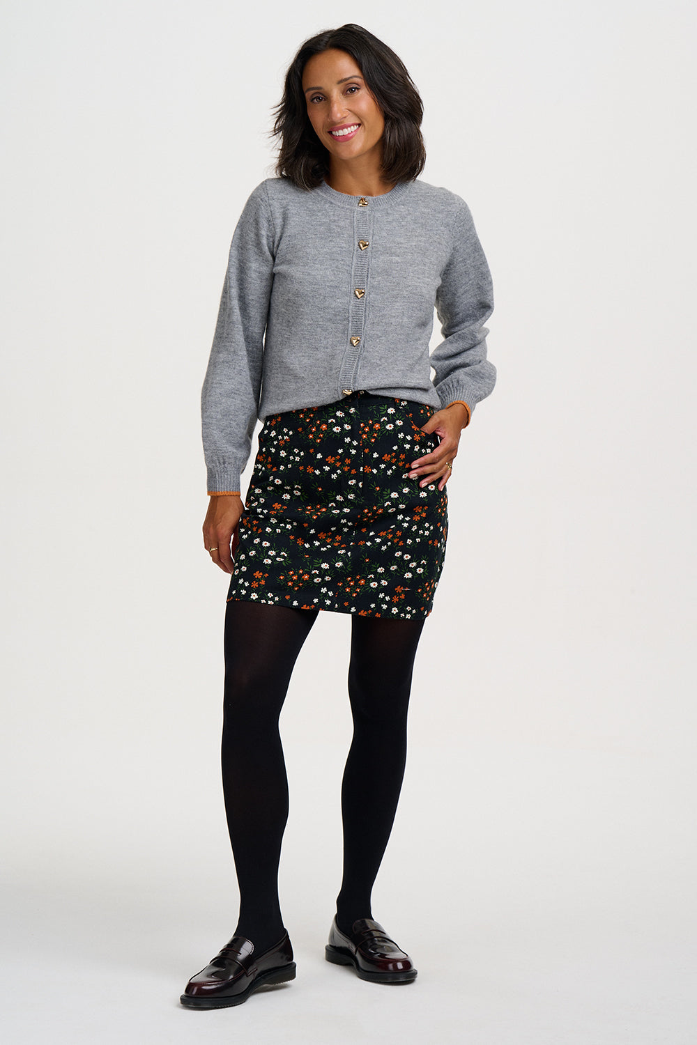 A woman modeling a light gray long-sleeve button-front cardigan, a black mini skirt with a colorful floral pattern, black tights, and black leather shoes against a plain white studio background.