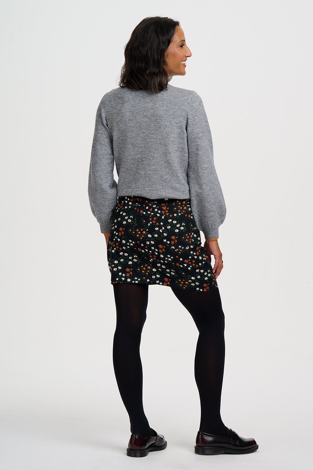 A woman is shown from the back wearing a heather gray long-sleeve knit sweater, a black mini skirt with a colorful floral print, black tights, and dark brown leather shoes, against a plain light gray studio background.
