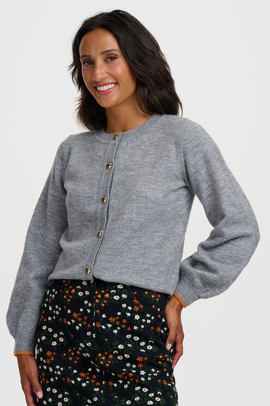 A woman modeling a light gray cropped button-front cardigan with long sleeves, paired with a black floral mini skirt, against a plain light gray studio background.