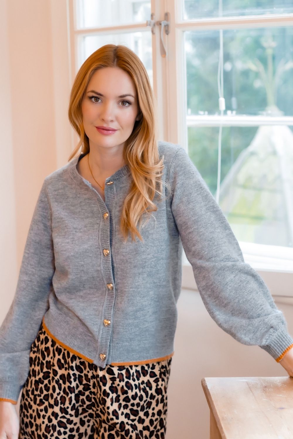 A woman wearing a light gray knit cardigan with gold buttons and leopard print trousers, standing in a brightly lit room with a window behind her.