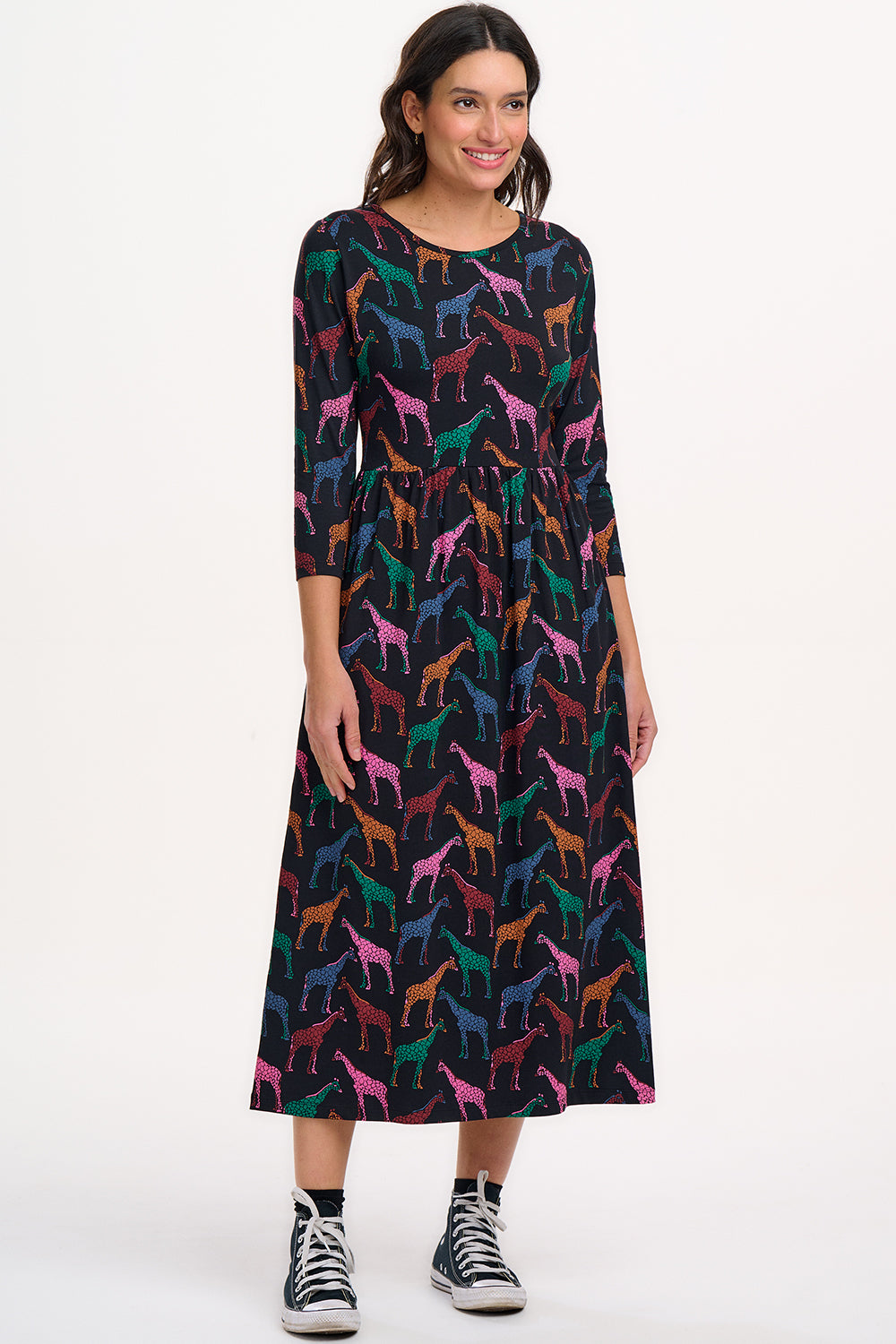 A model wearing a midi jersey dress with a gathered waist and 3/4 length sleeves. The dress has a black base with a colorful giraffe print.