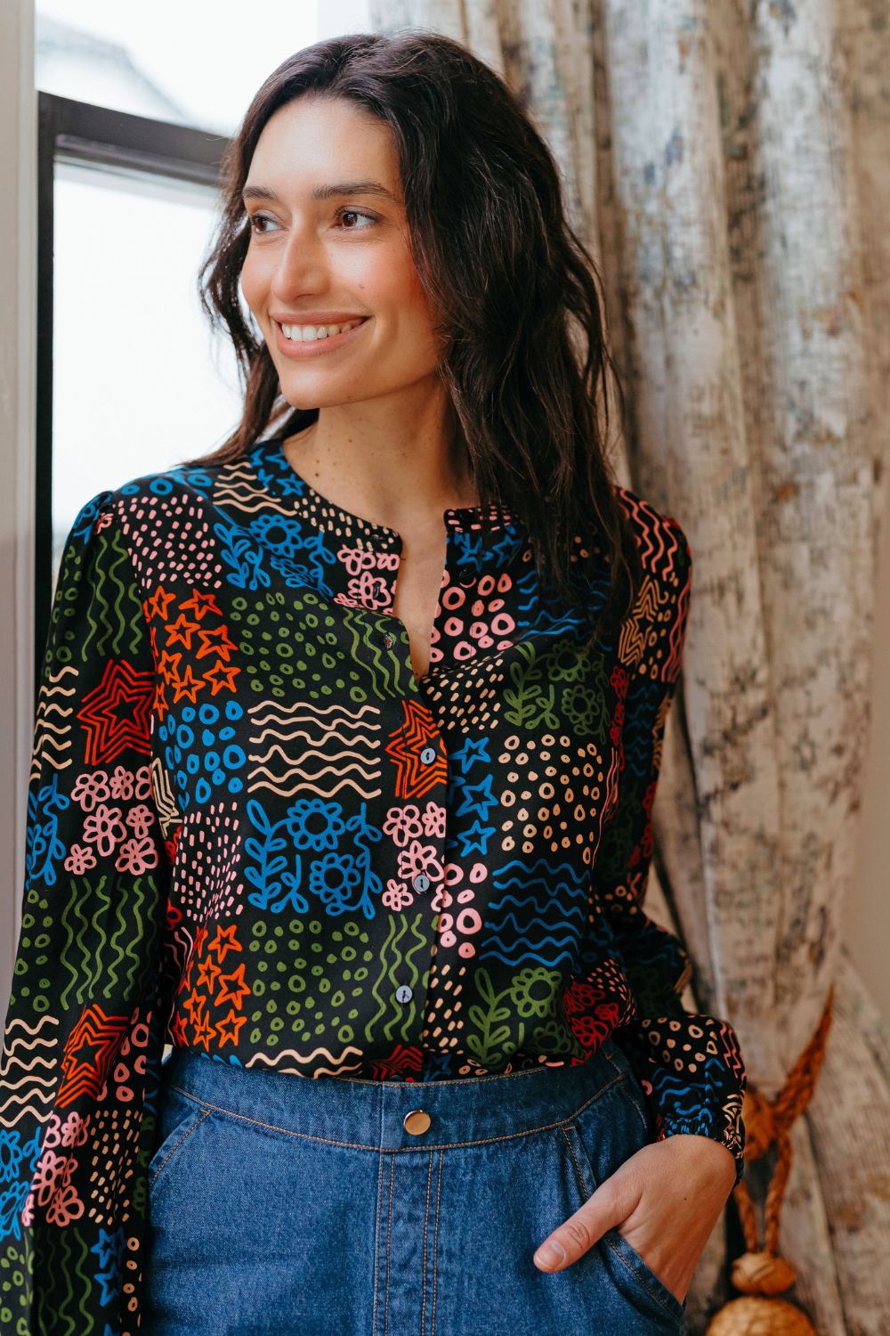 Woman wearing a colorful patterned blouse with blue pants indoors.