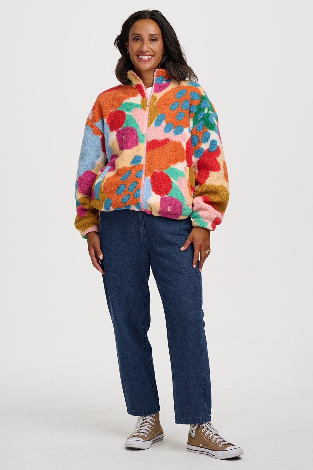 A woman modeling a colorful, fuzzy quarter-zip fleece jacket with a vibrant abstract floral pattern in orange, pink, blue, green, and yellow, paired with dark blue jeans and brown sneakers against a light gray studio background.