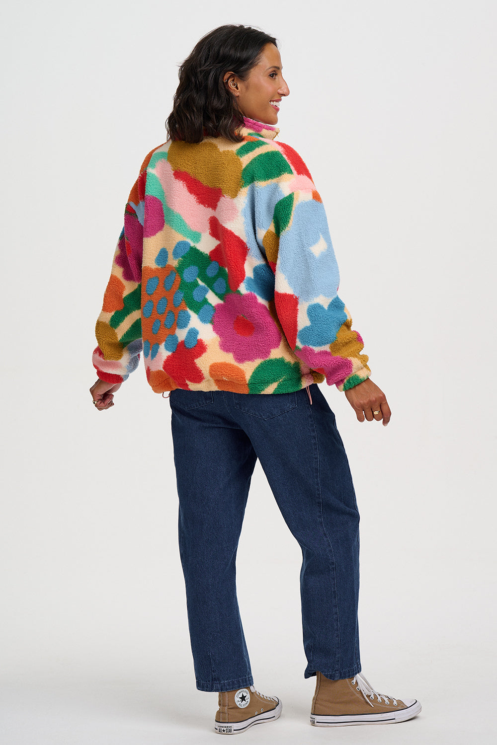 A woman wearing a colorful, long-sleeve fuzzy fleece pullover with a vibrant abstract floral pattern, paired with dark blue jeans and brown canvas sneakers, shown from a side-back angle against a light gray studio background.