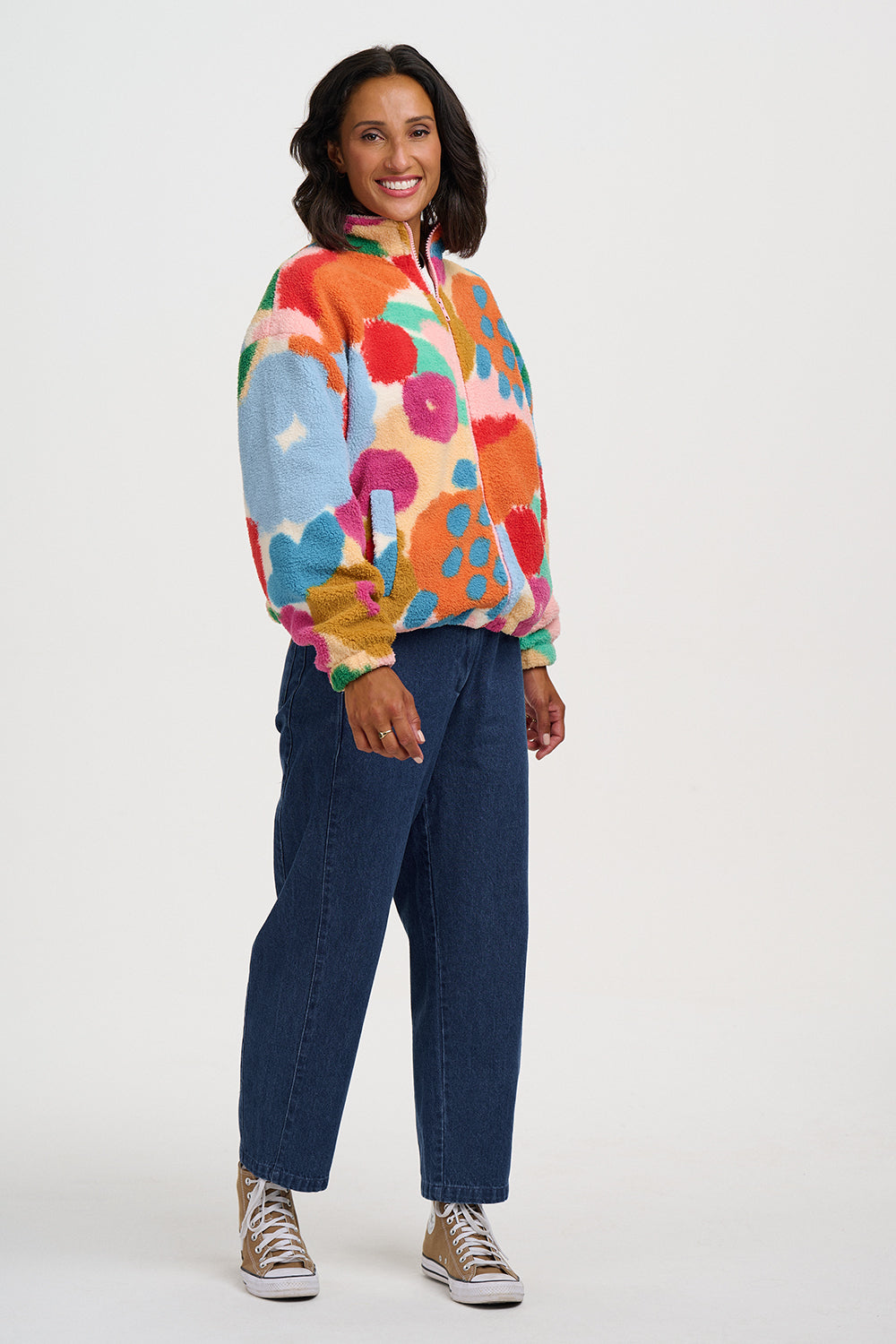 A woman modeling a colorful floral print fleece pullover jacket with blue jeans and brown sneakers, standing against a plain light gray studio background.