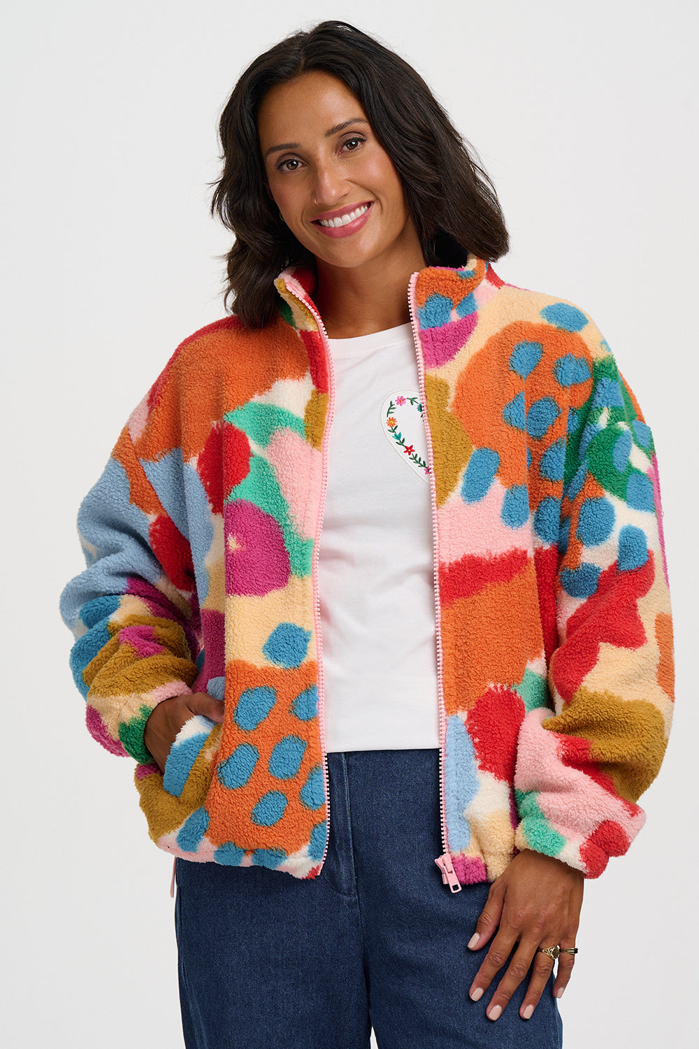 A woman wearing a colorful fuzzy fleece zip-up jacket with a multicolor abstract pattern, styled over a white t-shirt and blue jeans against a light gray background.