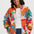 A woman wearing a colorful fuzzy fleece zip-up jacket with a multicolor abstract pattern, styled over a white t-shirt and blue jeans against a light gray background.