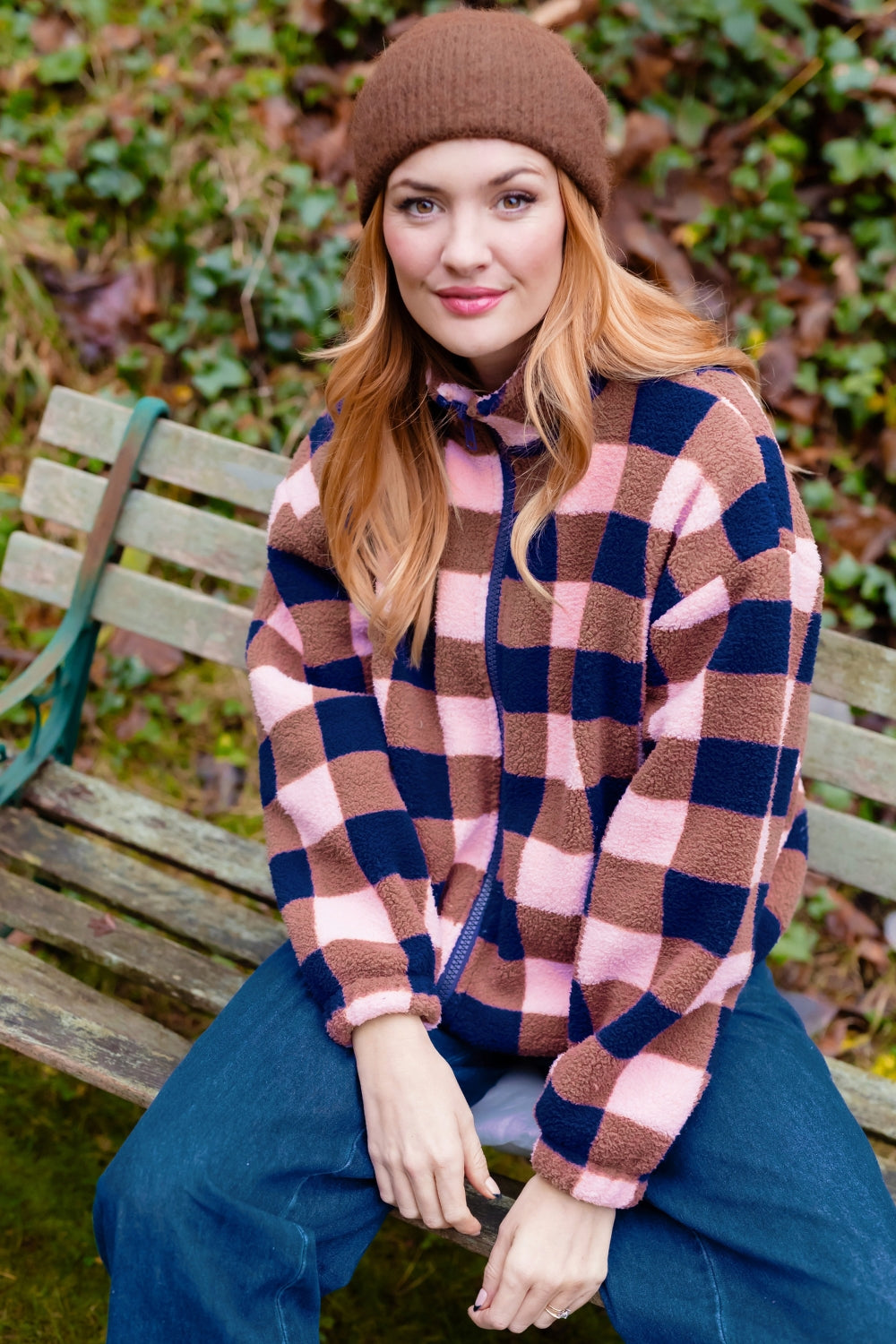A woman sitting on a wooden bench outdoors wearing a brown knit beanie, a pink and navy blue checkered fleece zip-up hoodie, and blue jeans.
