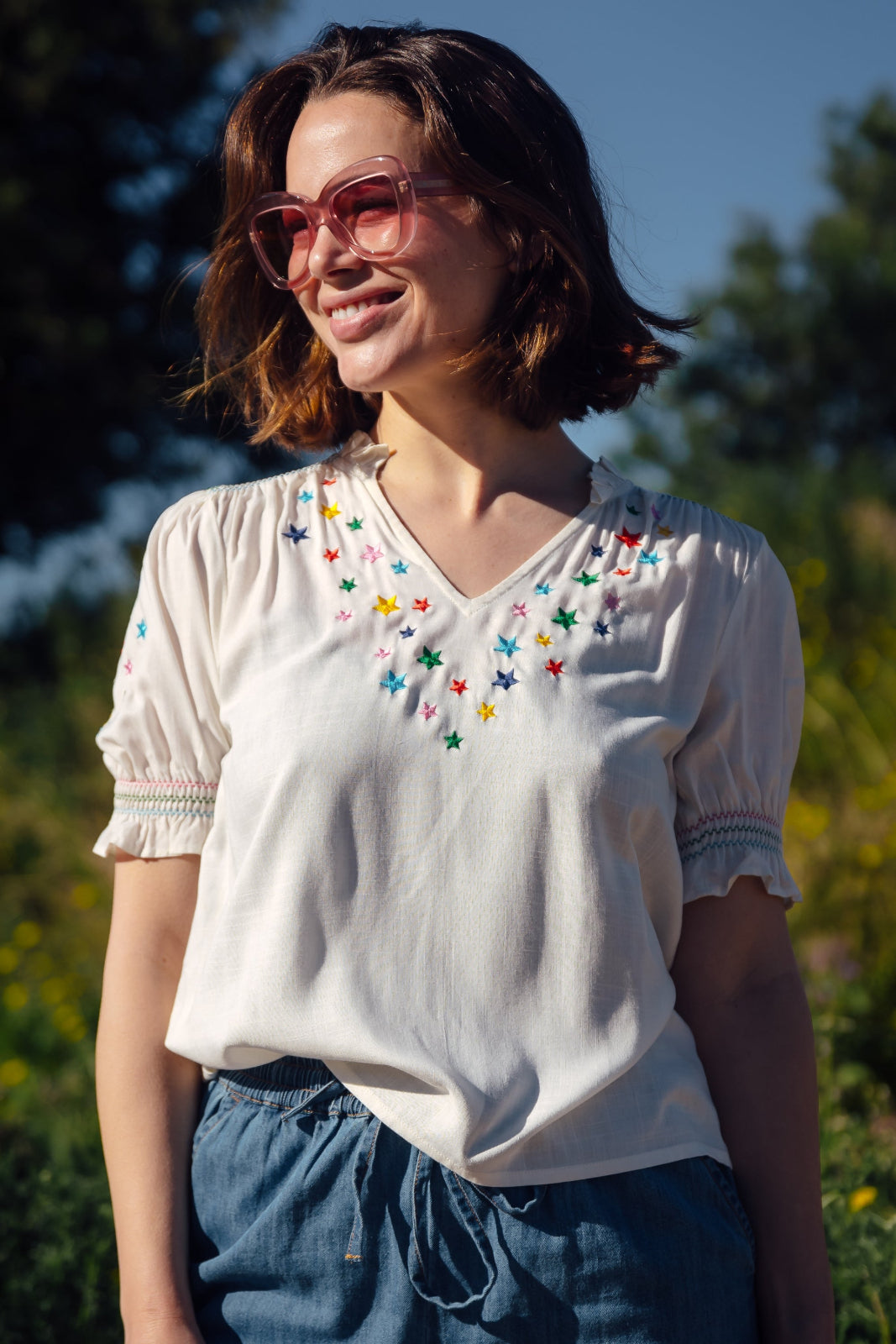 A woman smiling and wearing an off-white blouse with rainbow star embroidery around the neckline and on the sleeves, with a frill collar and shirred frill cuffs.