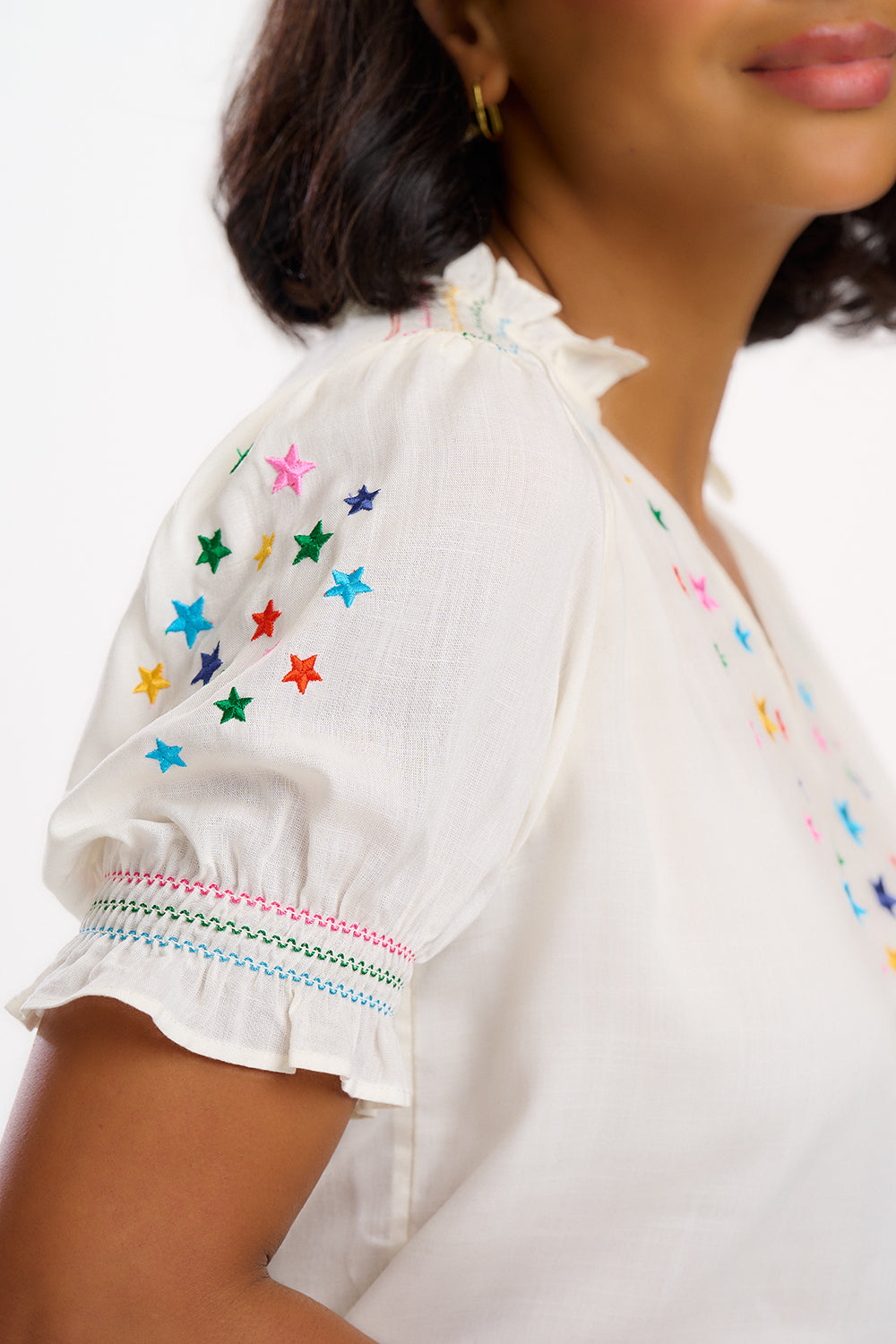 A woman modeling an off-white blouse with rainbow star embroidery around the neckline and on the sleeves, featuring a shirred frill cuff and a V-neckline.