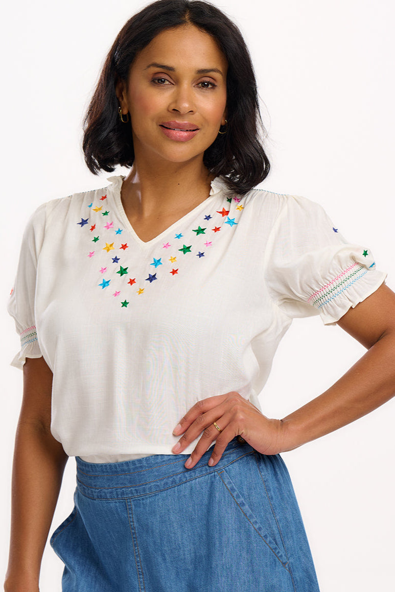 A woman modeling an off-white top with rainbow stars embroidery around the neckline and on the sleeves, a frill collar, and shirred frill cuffs.