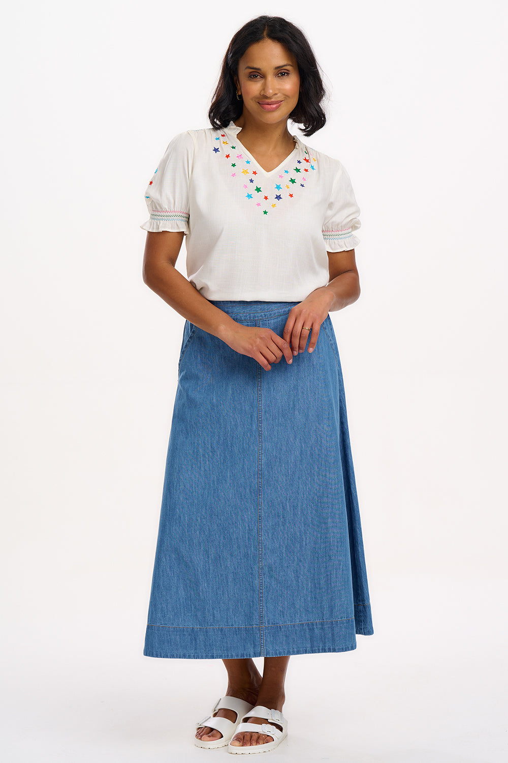 A woman modeling an off-white blouse with rainbow star embroidery around the neckline and on the sleeves, featuring a shirred frill cuff and a V-neckline.