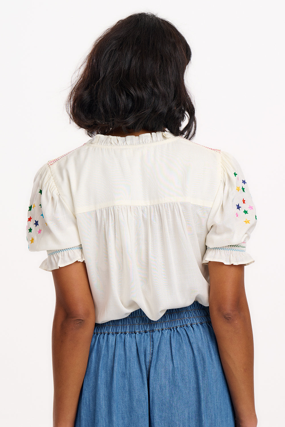 A woman modeling an off-white blouse with rainbow star embroidery around the neckline and on the sleeves, featuring a shirred frill cuff and a V-neckline.