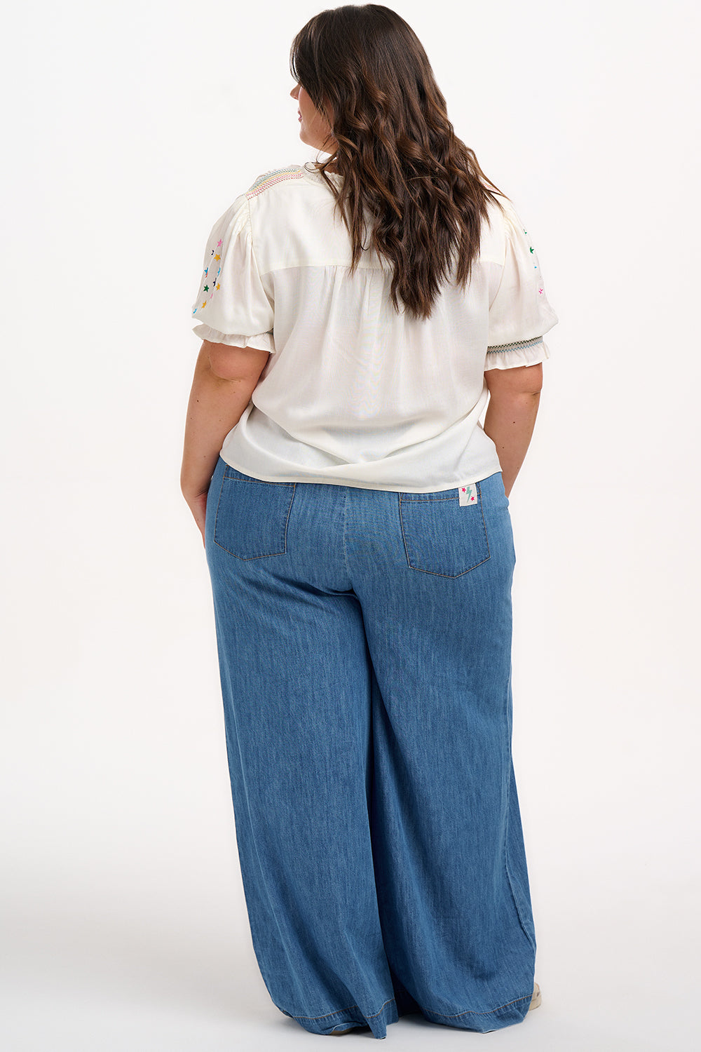 A woman modeling an off-white blouse with rainbow star embroidery around the neckline and on the sleeves, featuring a shirred frill cuff and a V-neckline.
