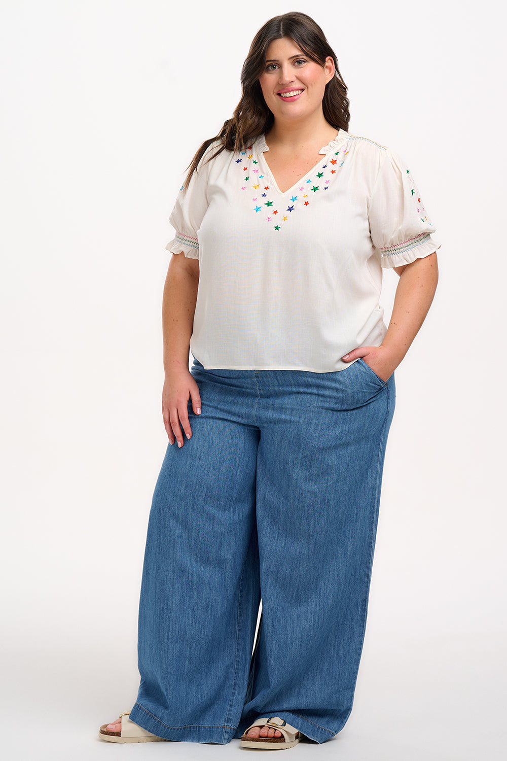 A woman modeling an off-white blouse with rainbow star embroidery around the neckline and on the sleeves, featuring a shirred frill cuff and a V-neckline.