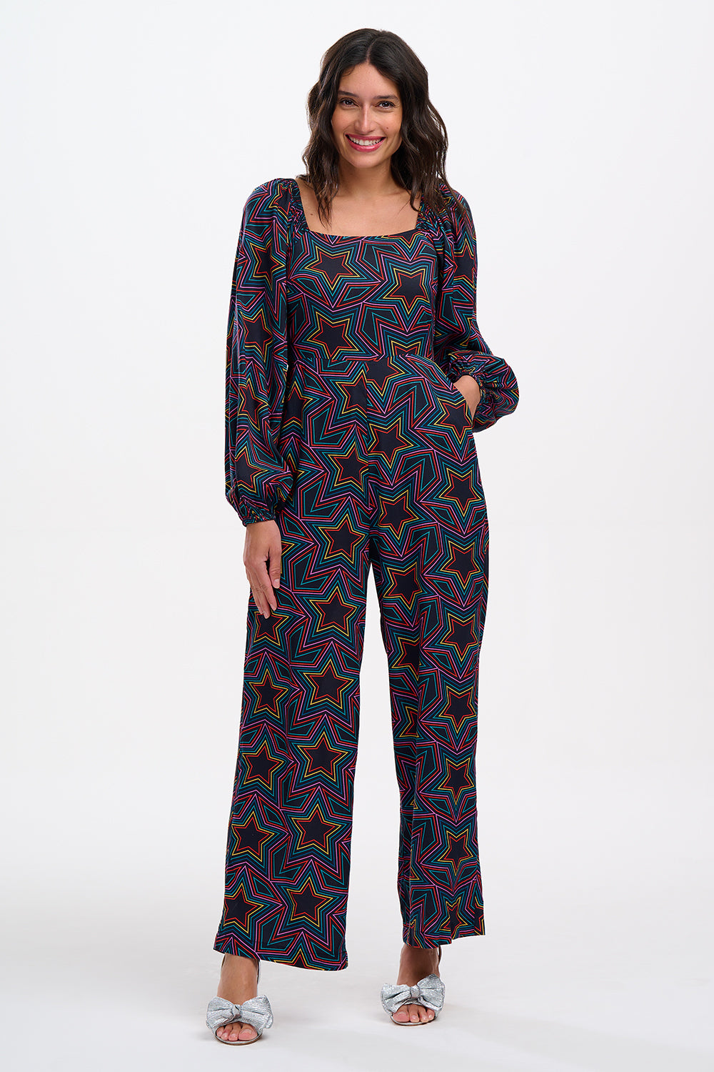 A model is standing and smiling, wearing a black and rainbow star explosion patterned jumpsuit with a square neckline, long blouson sleeves, a fitted waist, and wide-leg trousers.