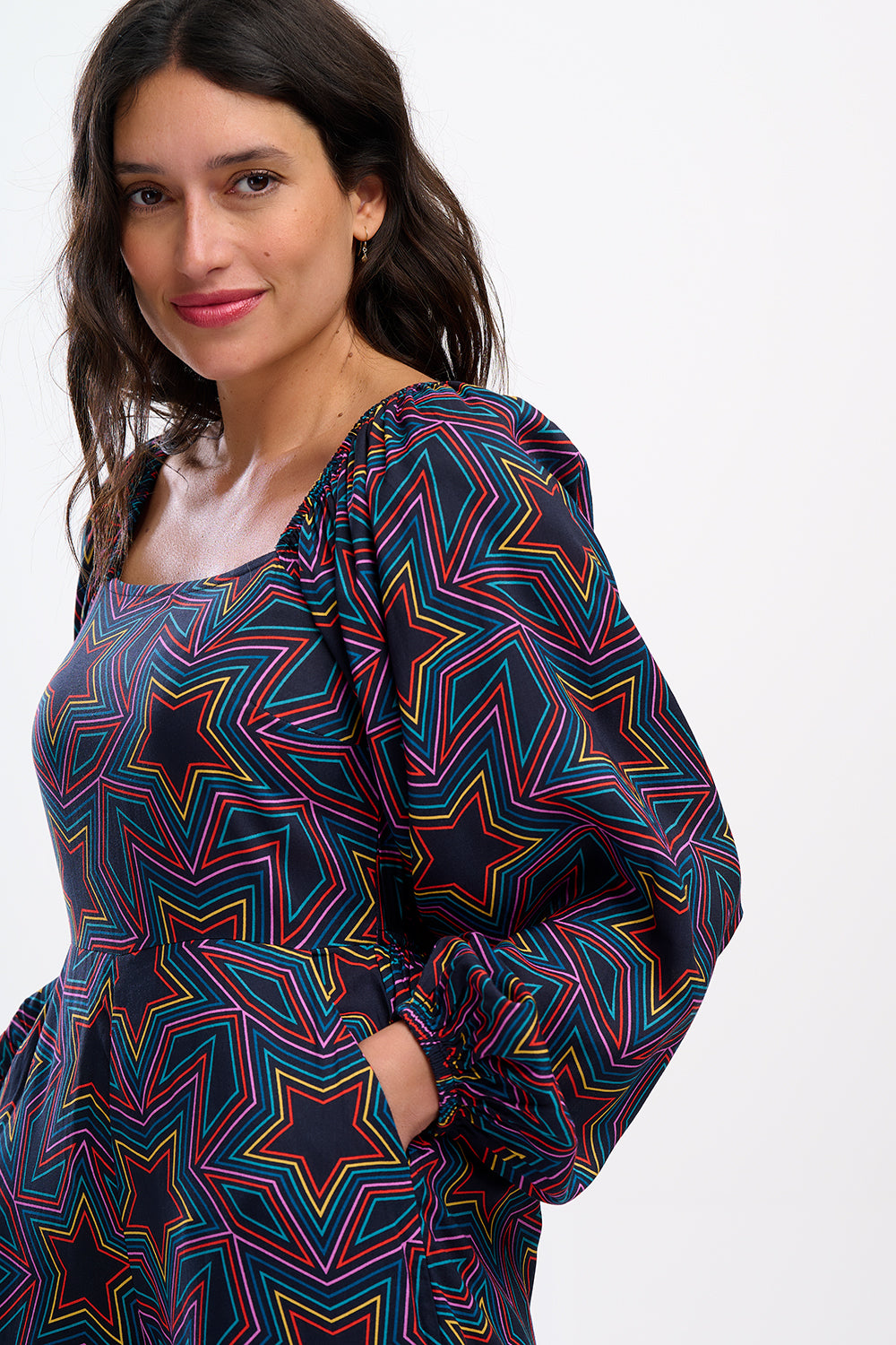 A model is standing and smiling, wearing a black and rainbow star explosion patterned jumpsuit with a square neckline, long blouson sleeves, a fitted waist, and wide-leg trousers.