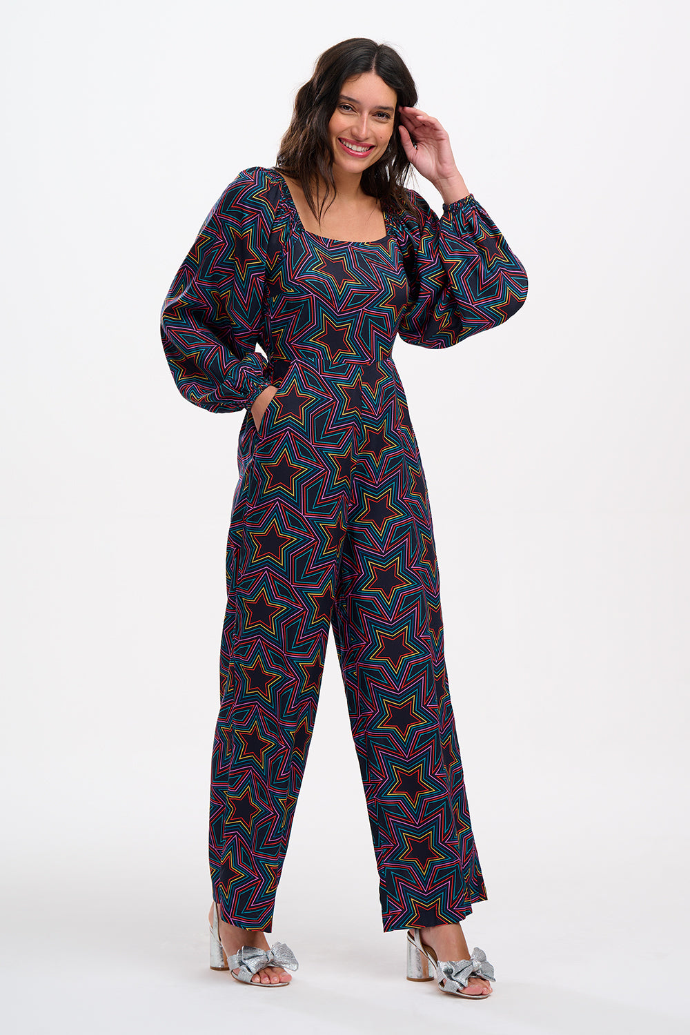 A model is standing and smiling, wearing a black and rainbow star explosion patterned jumpsuit with a square neckline, long blouson sleeves, a fitted waist, and wide-leg trousers.
