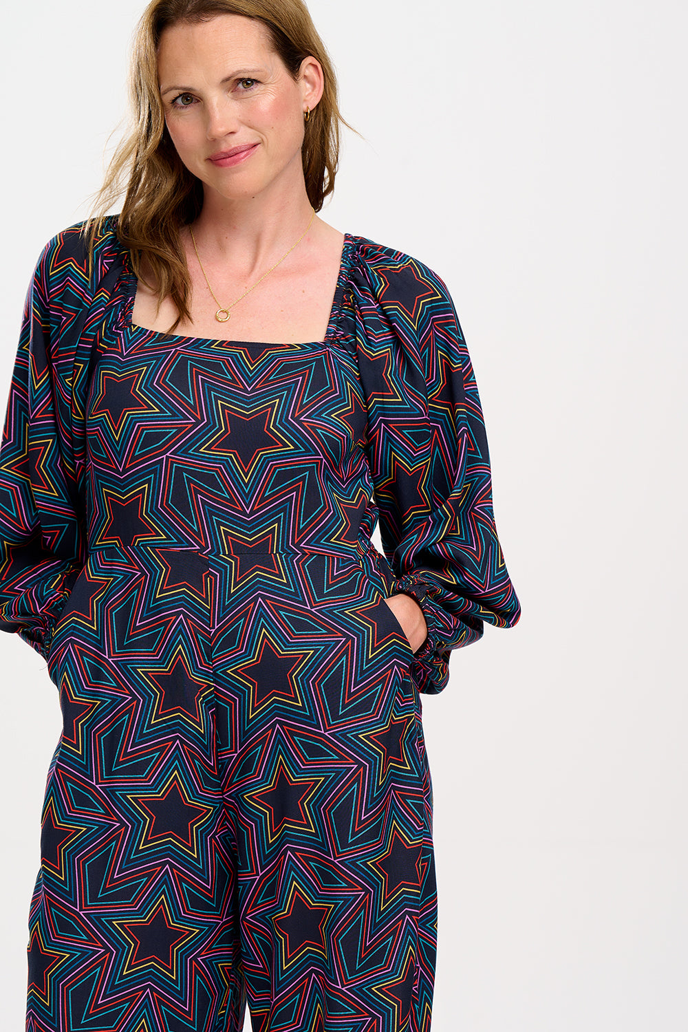 A model is standing and smiling, wearing a black and rainbow star explosion patterned jumpsuit with a square neckline, long blouson sleeves, a fitted waist, and wide-leg trousers.